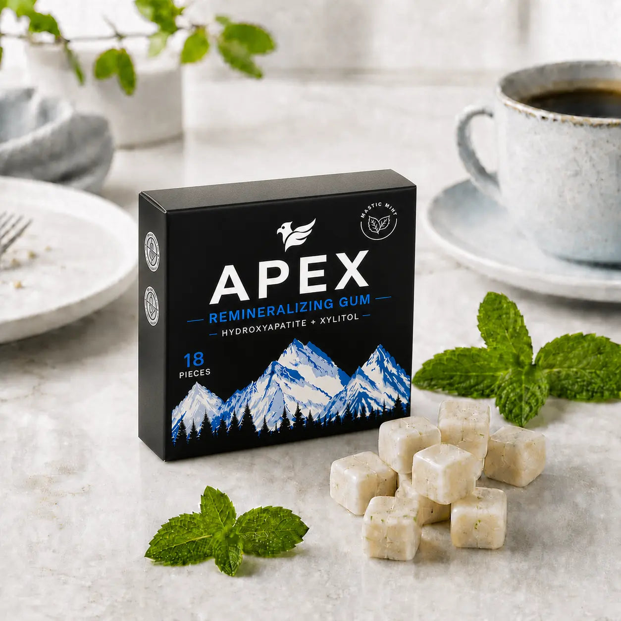 A box of APEX Remineralizing Gum sits on a light stone surface with several pale gum cubes in front, mint leaves around it, and a cup of coffee in the background.