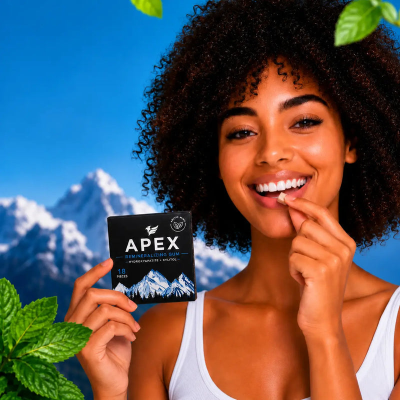 A smiling woman with dark curly hair holds a black APEX toothpaste packet in one hand and applies toothpaste to her teeth with the other. She is set against a blue sky and snow-capped mountain background with green leaves in the corners.