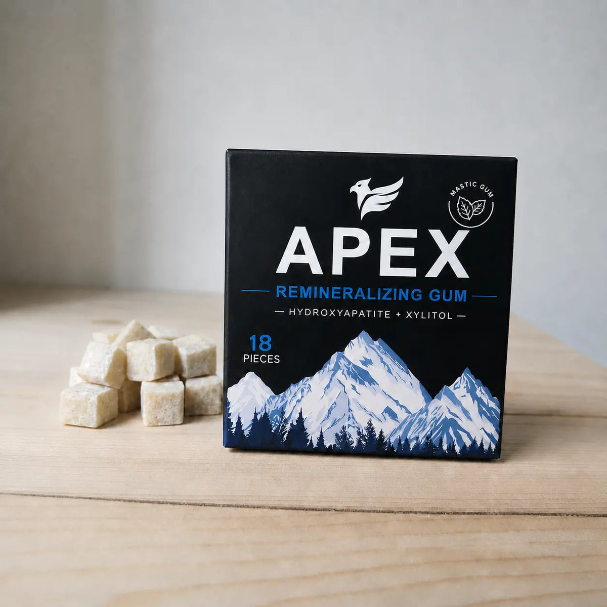 A black box of APEX Mastic Mint Chewing Gum with a blue and white mountain illustration, set on a light wooden surface next to several beige gum cubes.