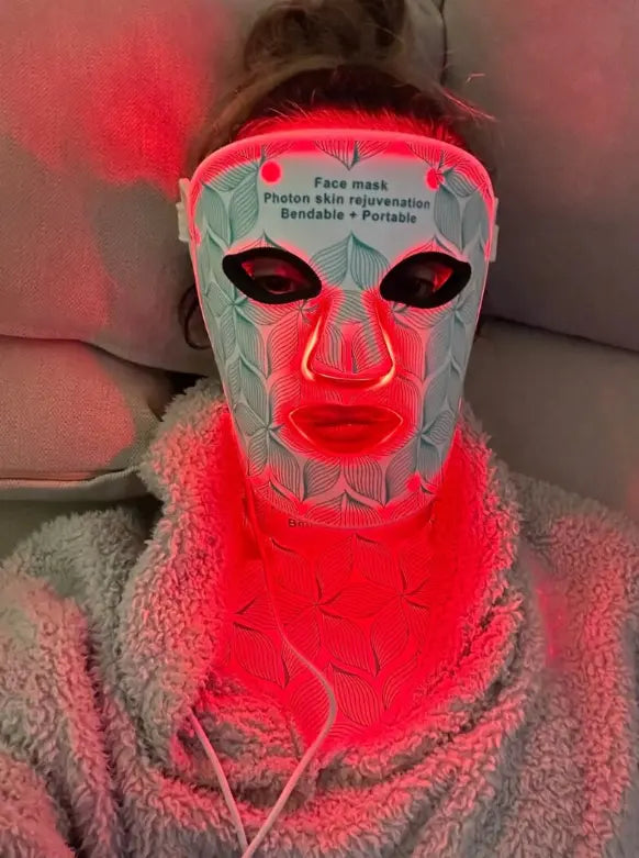 Woman wearing a flexible LED face and neck light therapy mask emitting red light during a home skincare treatment session.